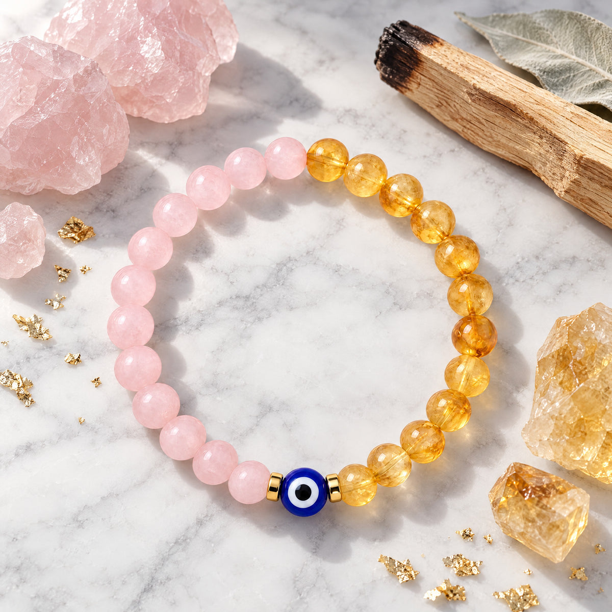 Rose quartz and citrine evil eye bracelet on marble with rose quartz pieces, citrine stones, sage, wood, and gold flakes nearby.